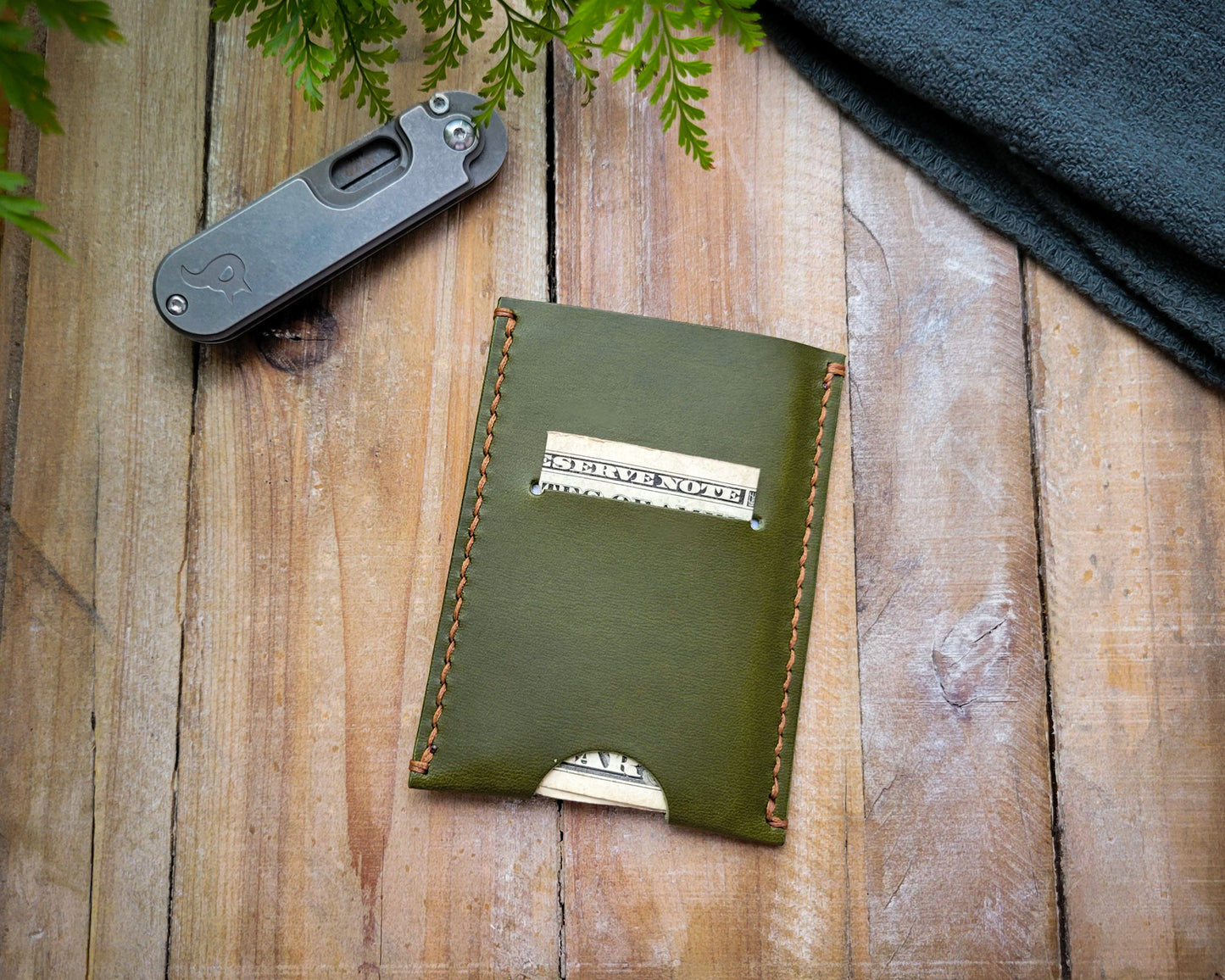 A pine green ultralight wallet made of Italian full grain leather, displayed on a wooden surface, with a knife and foliage in the background.