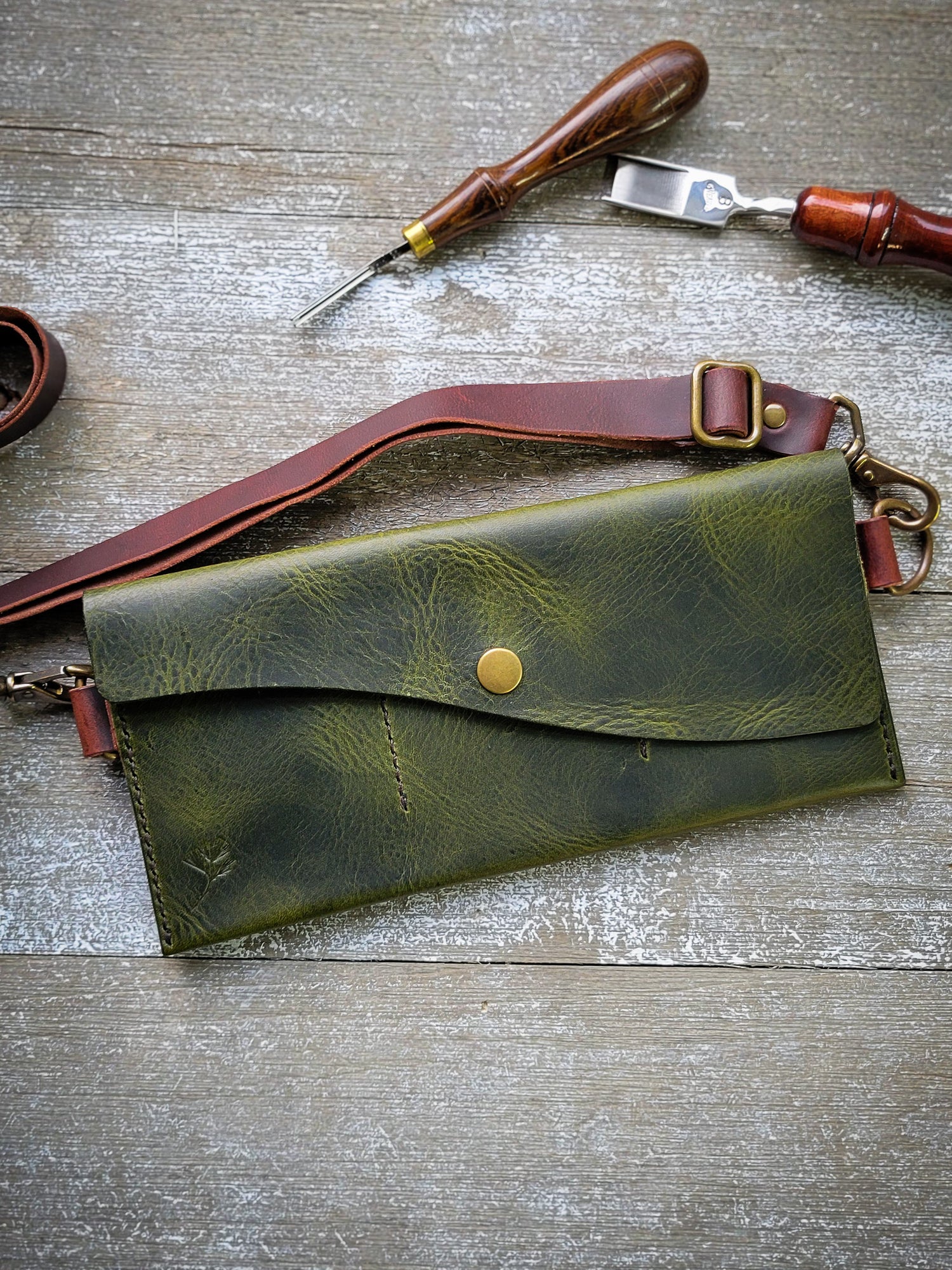 Green leather clutch bag with brass clasp on a wooden surface with leather tools.