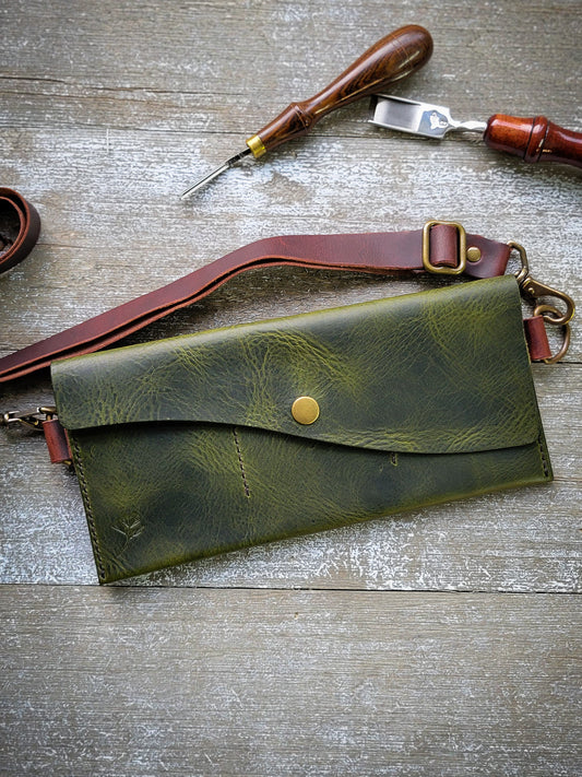 Green leather clutch bag with brass clasp on a wooden surface with leather tools.