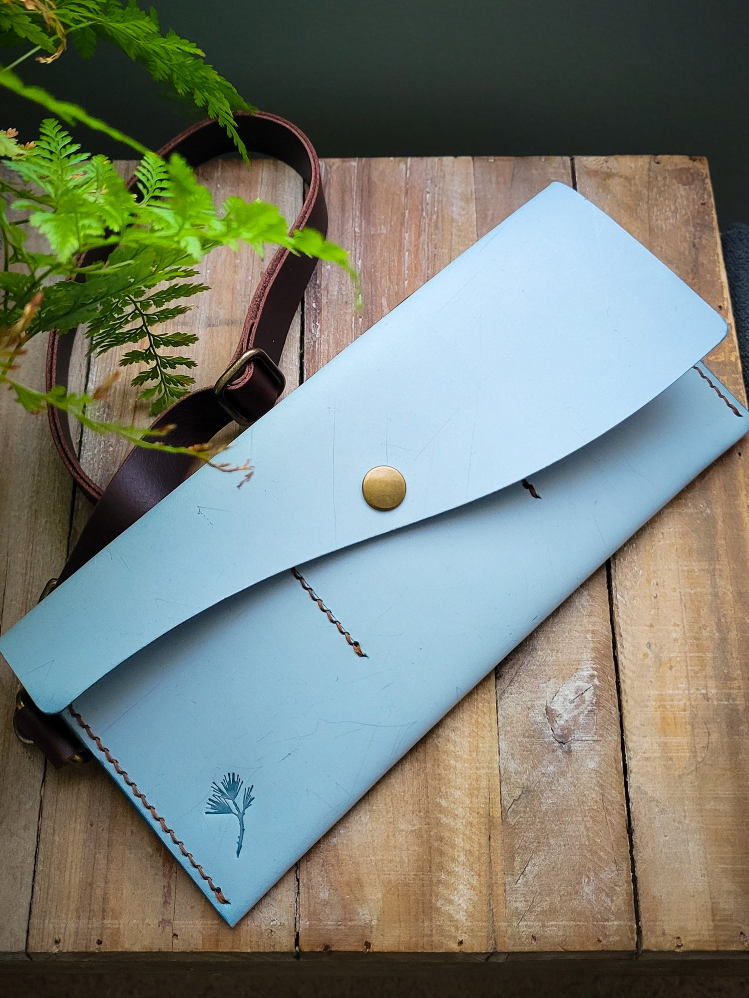 Light blue leather pouch with a button on a wooden surface with a plant in the background
