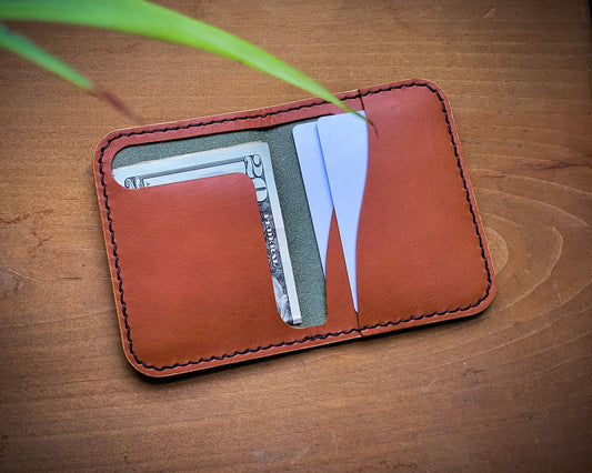 Green Pueblo Leather minimalist wallet with Tan Wickett & Craig interior laying open on top of distressed wood. It is holding folded cash and a couple cards. 