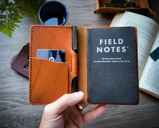 Tan Wickett and Craig field notes journal cover with red thread held open. A leather valet tray and open book in the background