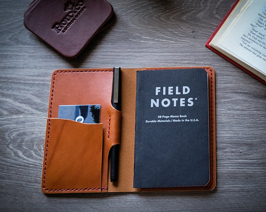 Tan Wickett and Craig field notes journal cover with red thread laying open on a desk. Leather coasters and open book in the background