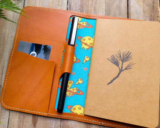 Tan leather notebook cover with pen holder and notebook. The inside is lined with fabric printed with planets made of pepperoni pizza on a turquoise background.