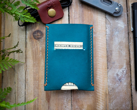 Turquoise Blue ultralight minimalist card wallet made from Minerva Italian full grain leather. It is sitting on a rustic wood table with headphones, pen, and pocket knife in the background.