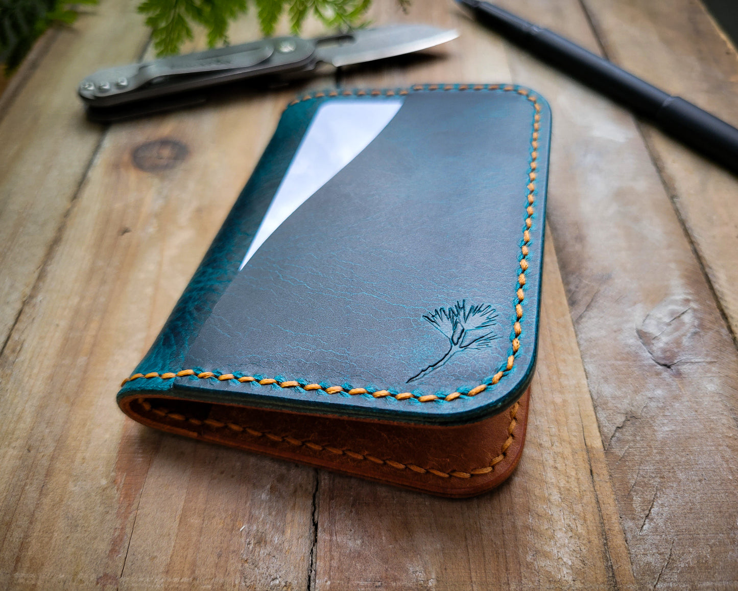 A close-up shot of the Rouxco white pine branch logo embossed on aTurquoise and orange Italian leather wallet sitting closed on a rustic table. A Black Fox pocket knife and a pen are in the background