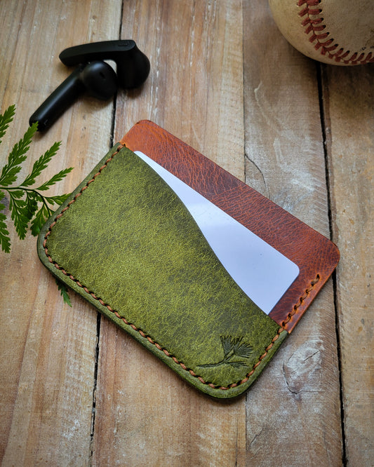 Green leather cardholder on a wooden surface with a baseball and tumbler in the background.