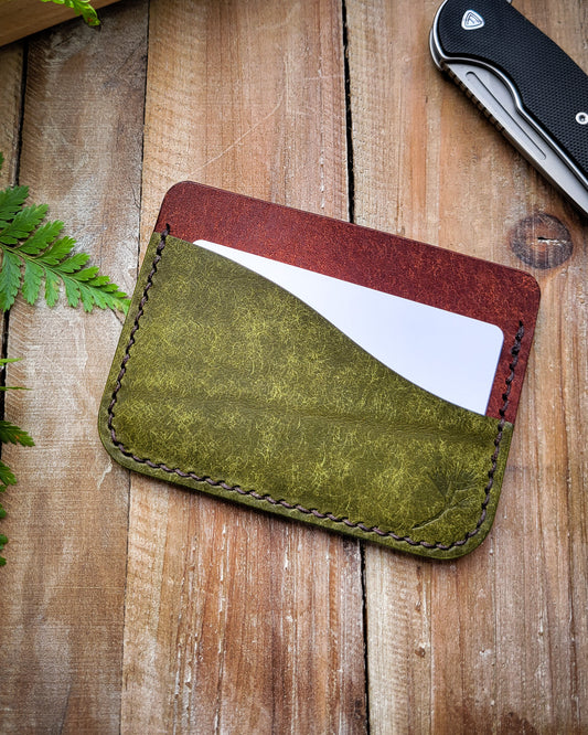Green leather cardholder with a white card on a wooden surface with a knife and plant.