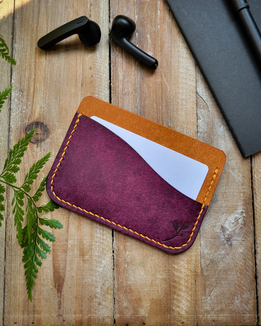 Purple leather cardholder with orange stitching on a wooden surface with black earbuds and a pen.