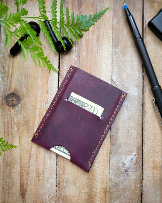 Purple leather wallet with money on a wooden surface with greenery and pens.