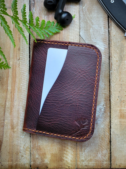 Brown leather cardholder with a white card on a wooden surface with a fern and black object in the background.