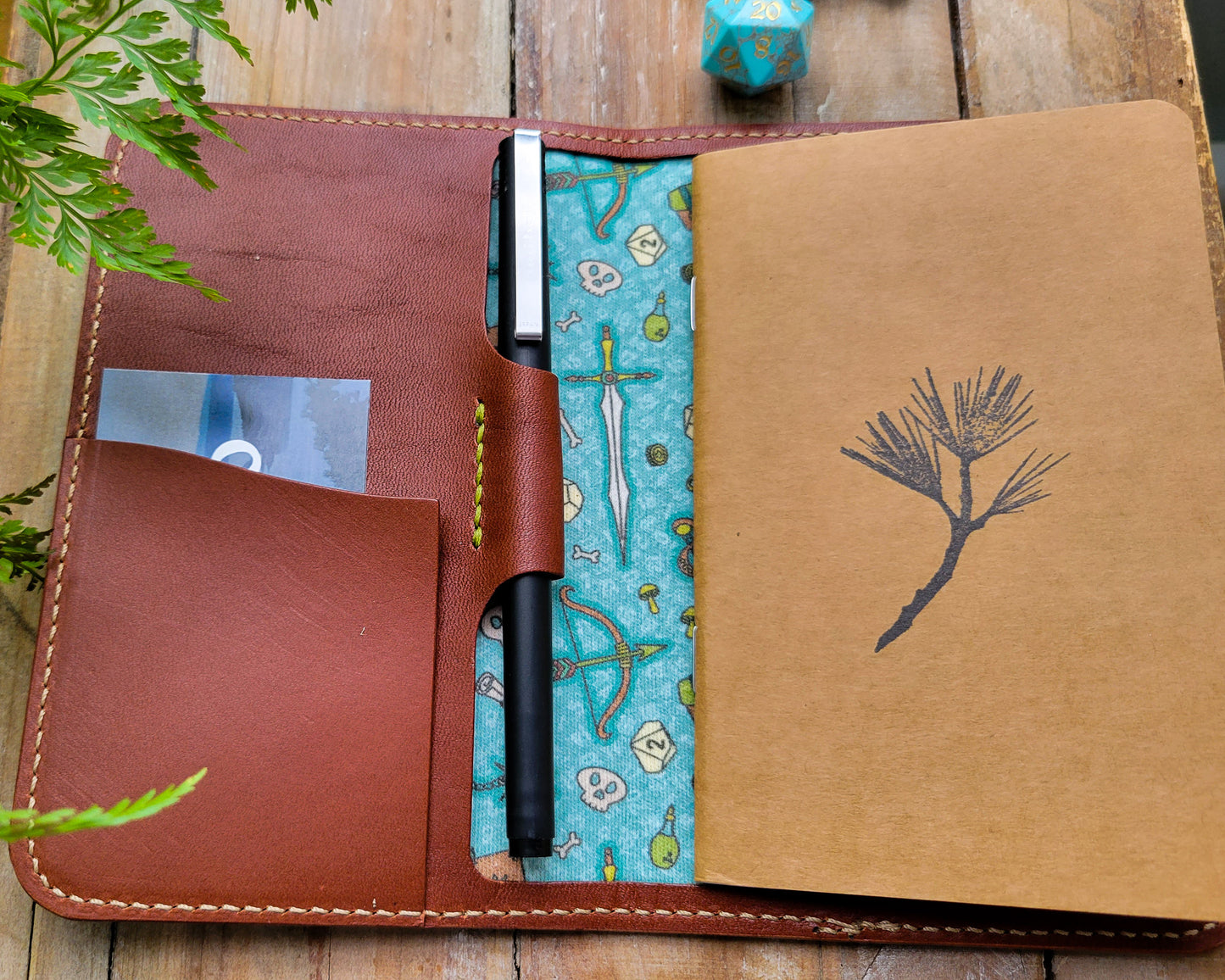 A brown leather notebook cover lined with D&D TTRPG fabric featuring a sword, bow, and dice. 