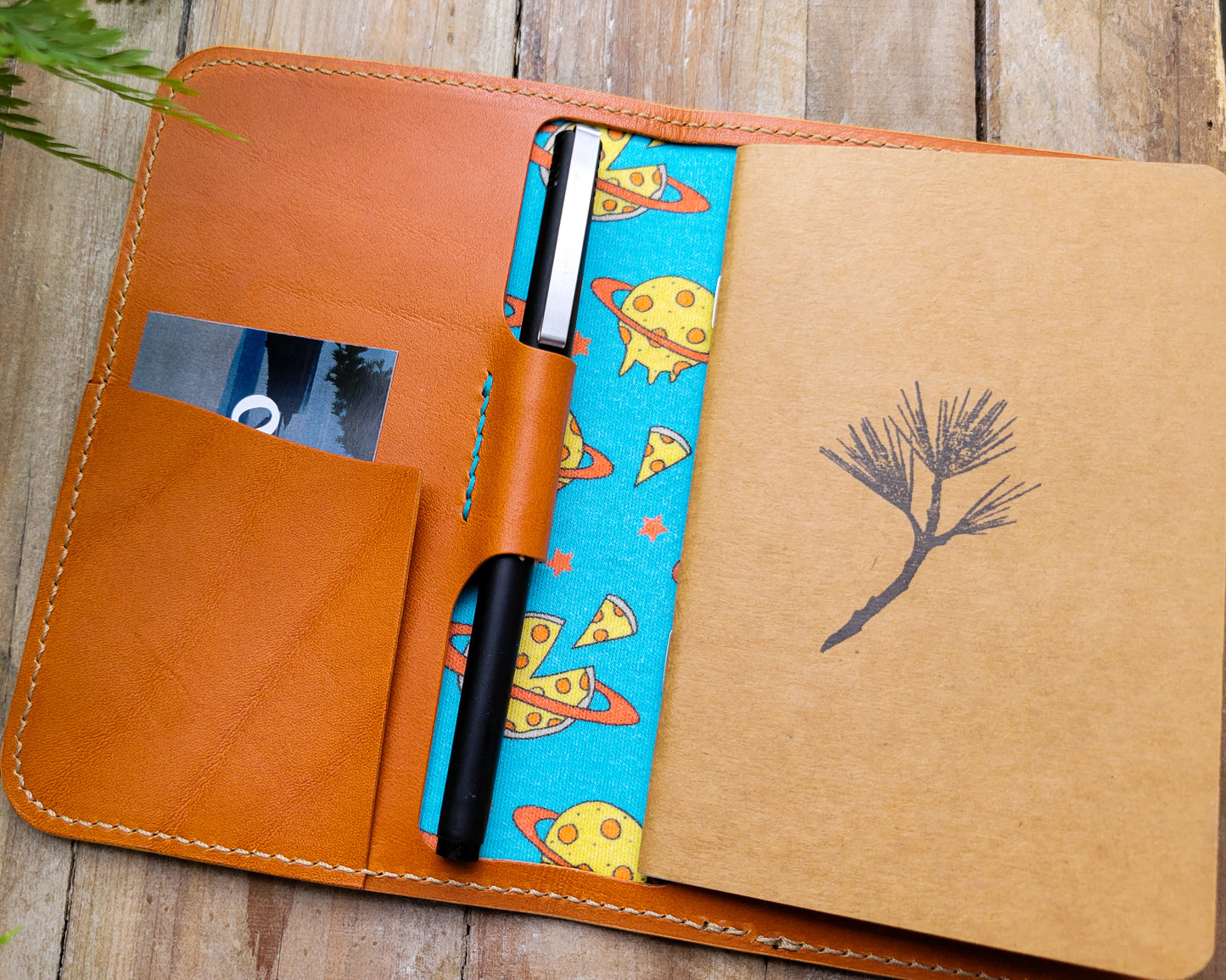 Tan leather notebook cover with pen holder and notebook. The inside is lined with fabric printed with planets made of pepperoni pizza on a turquoise background.