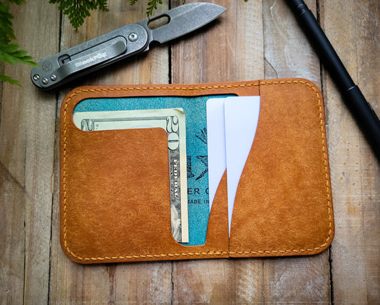 An Italian leather wallet sitting open on a rustic table. It has a cash flap on the left side for folded bills and two card slots on the right side.  A Black Fox pocket knife and a pen are in the background