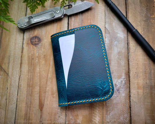 Turquoise and orange Italian leather wallet sitting closed on a rustic table. A Black Fox pocket knife and a pen are in the background